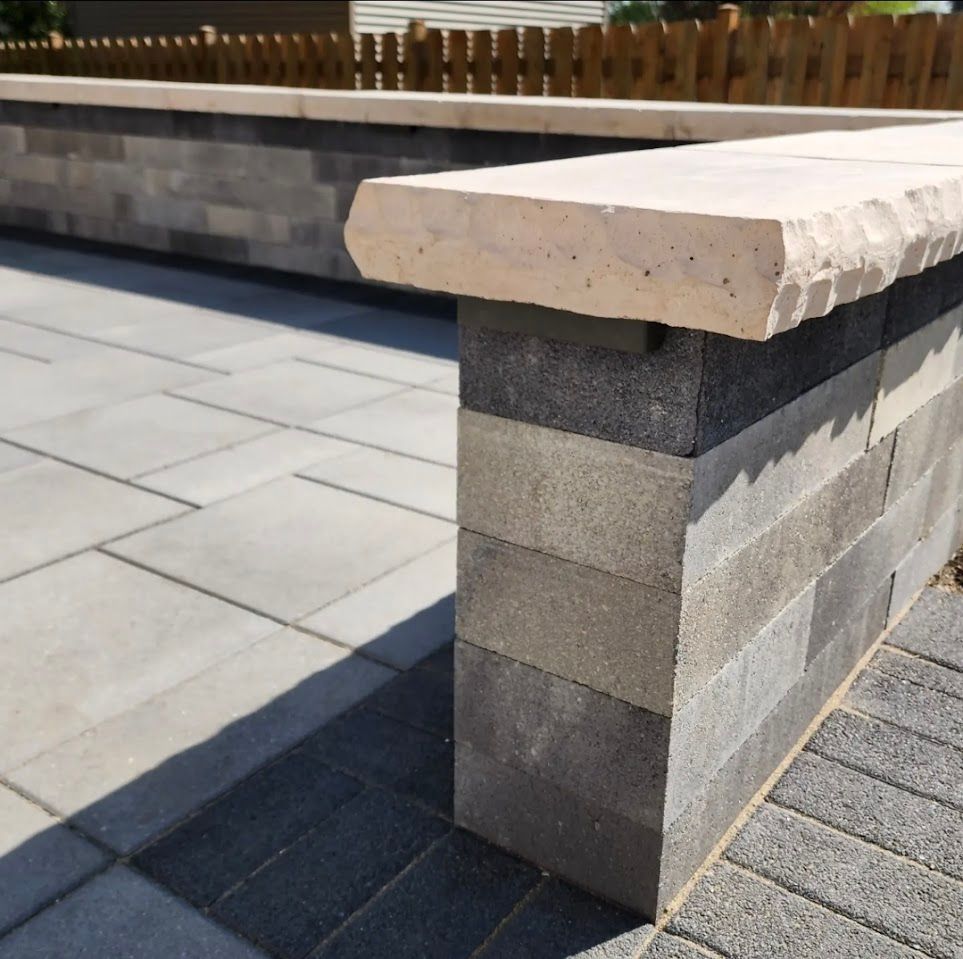 A low stone wall with a light-colored capstone bordering a grey paver patio and a wooden fence.