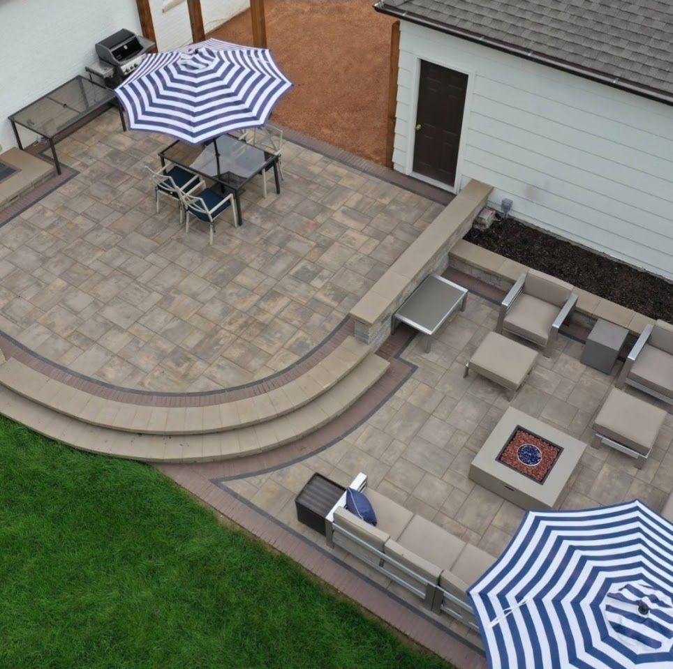 High-angle view of a backyard patio with pavers, two striped umbrellas, a dining set, fire pit, and sectional seating.