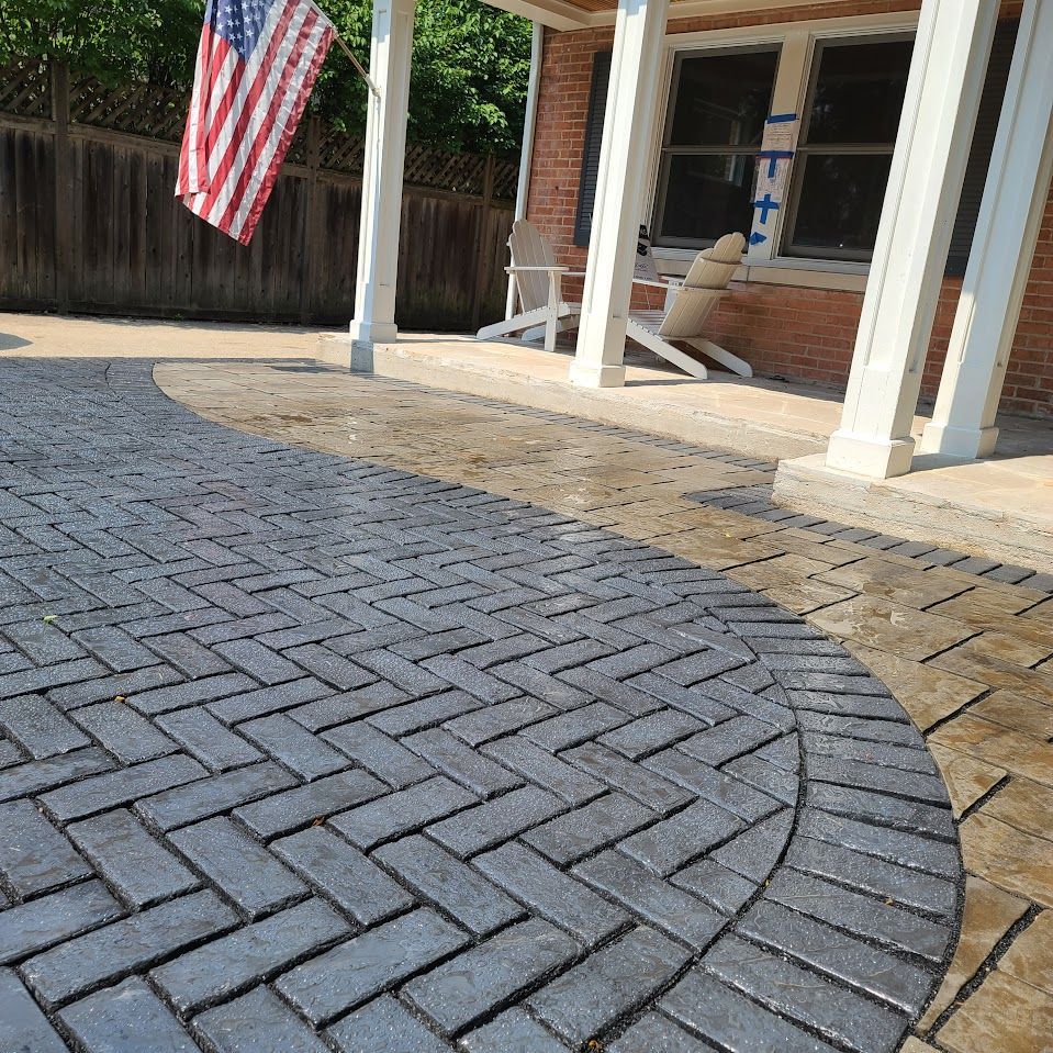 A brick-paved patio with a herringbone center pattern and a curved border next to a porch with columns and an American flag.