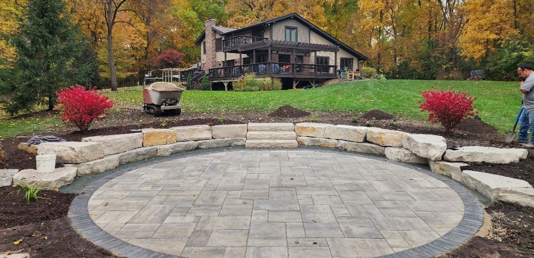 A circular stone patio with a retaining wall and central steps, set in a backyard near a house during autumn.
