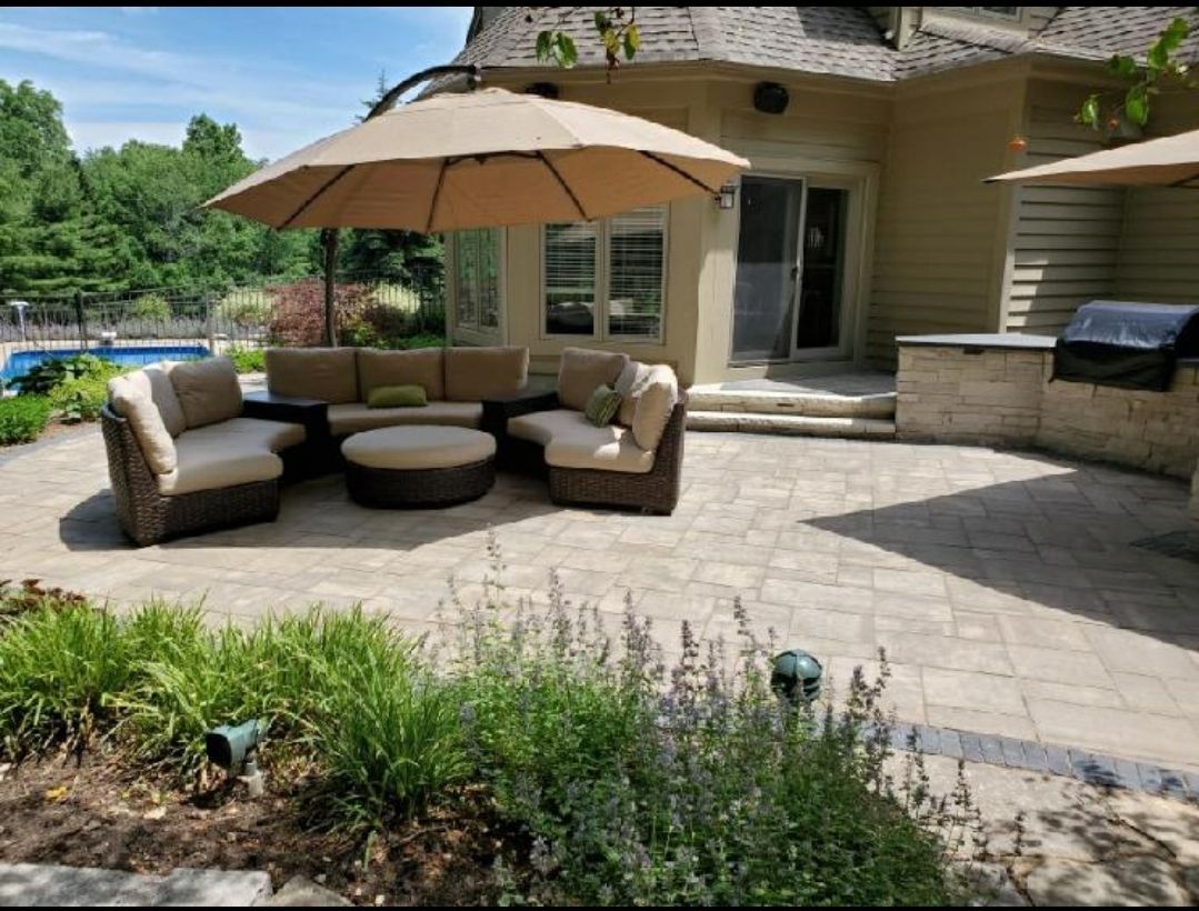 A paved patio with outdoor furniture under a beige umbrella, featuring a corner sofa, circular ottoman, and a grill area.