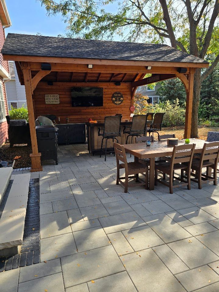 A wooden backyard pavilion with a flat-screen TV, a bar with high chairs, and a dining table on a stone patio.