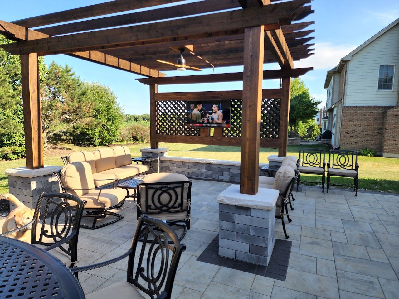 A patio features a wooden pergola with a mounted TV, cushioned seating, stone pillars, and a metal dining set.