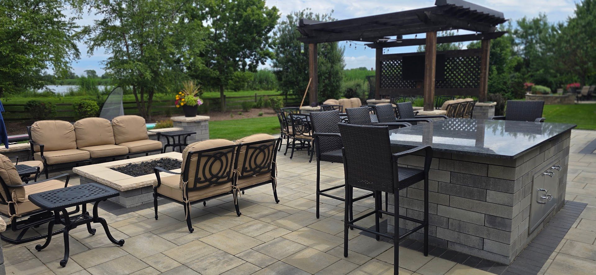 A luxury outdoor patio featuring a stone island bar, a fire pit with tan lounge seating, and a shaded pergola.