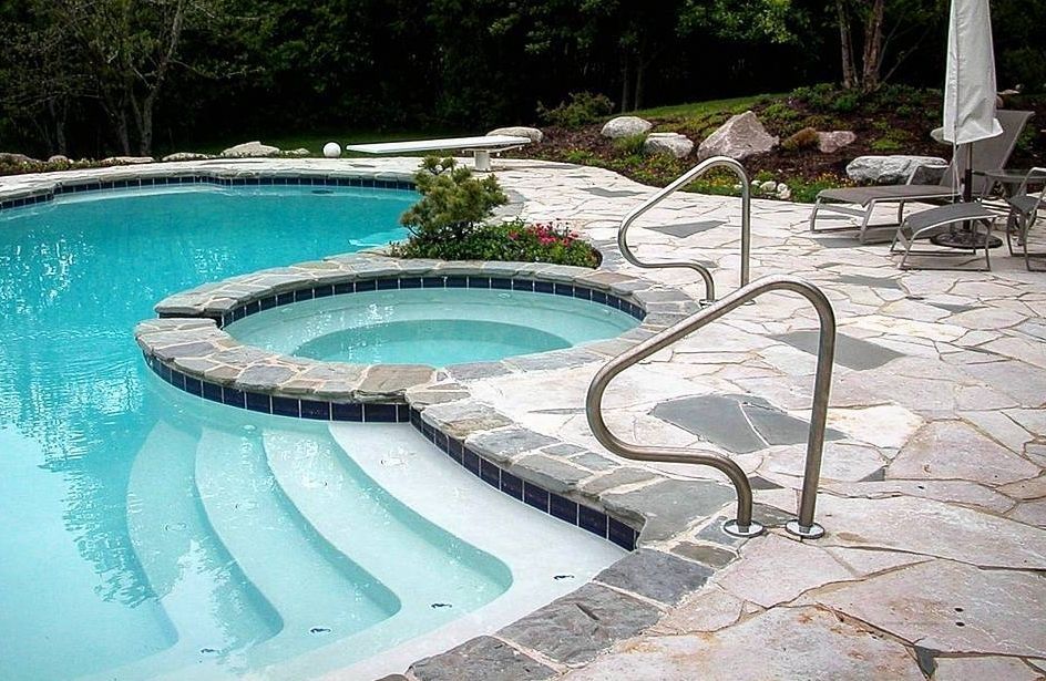 A turquoise pool with a raised circular spa, stone decking, and two stainless steel handrails for entry.