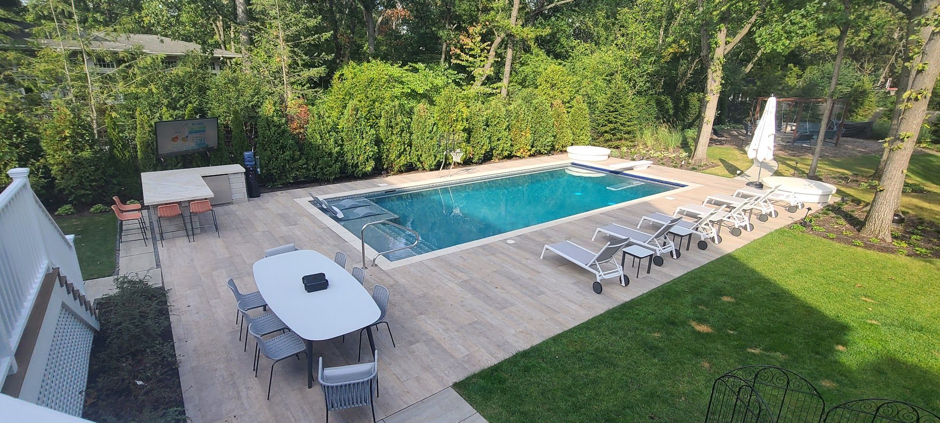 A rectangular swimming pool surrounded by a patio with lounge chairs and dining sets in a lush, tree-lined backyard.