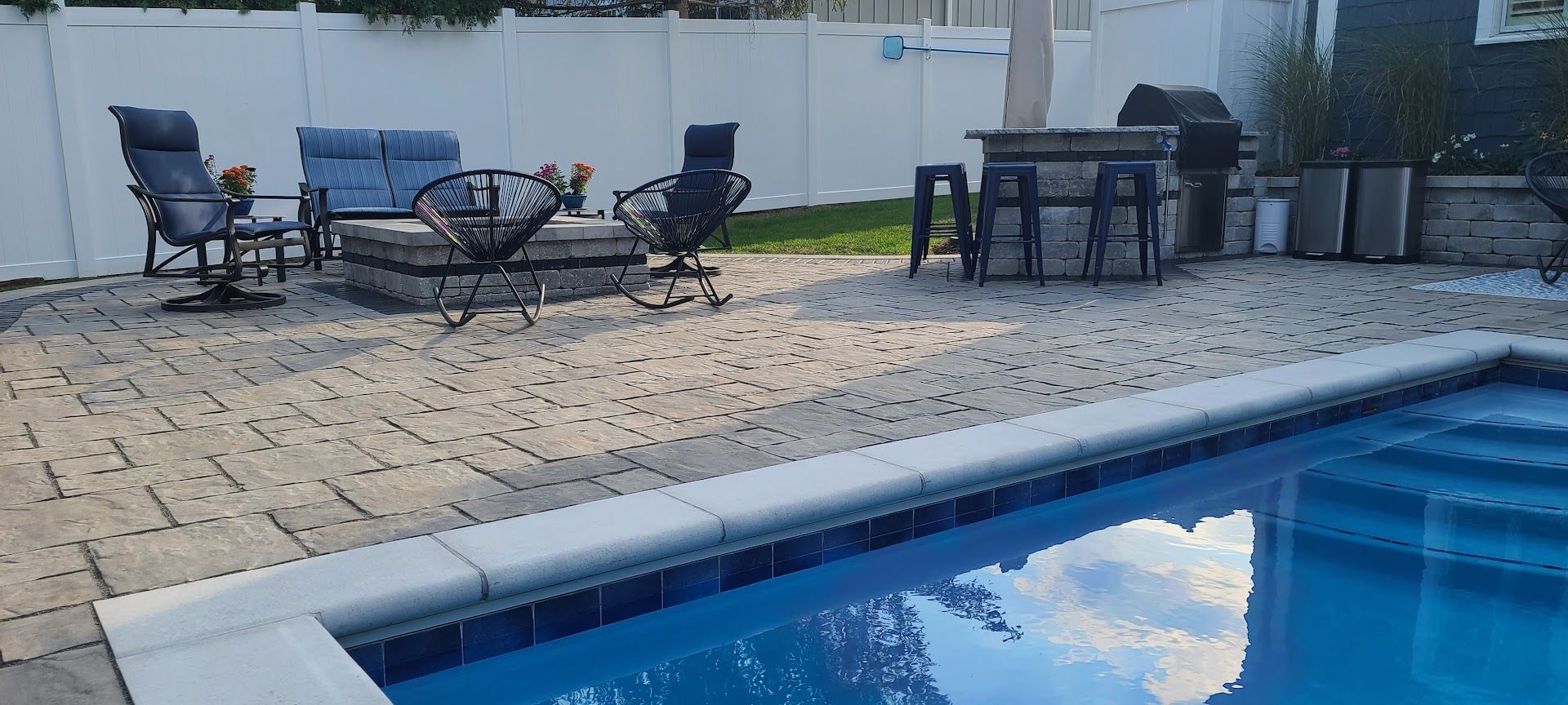 A backyard patio with a pool in the foreground, featuring a lounge area with seating, a fire pit, and a small outdoor bar.