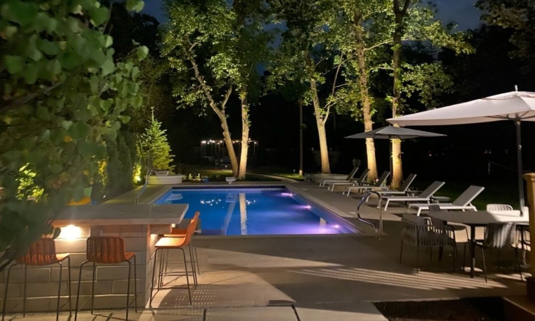 An illuminated backyard pool at night, featuring patio furniture, bar seating, and trees lit against a dark background.
