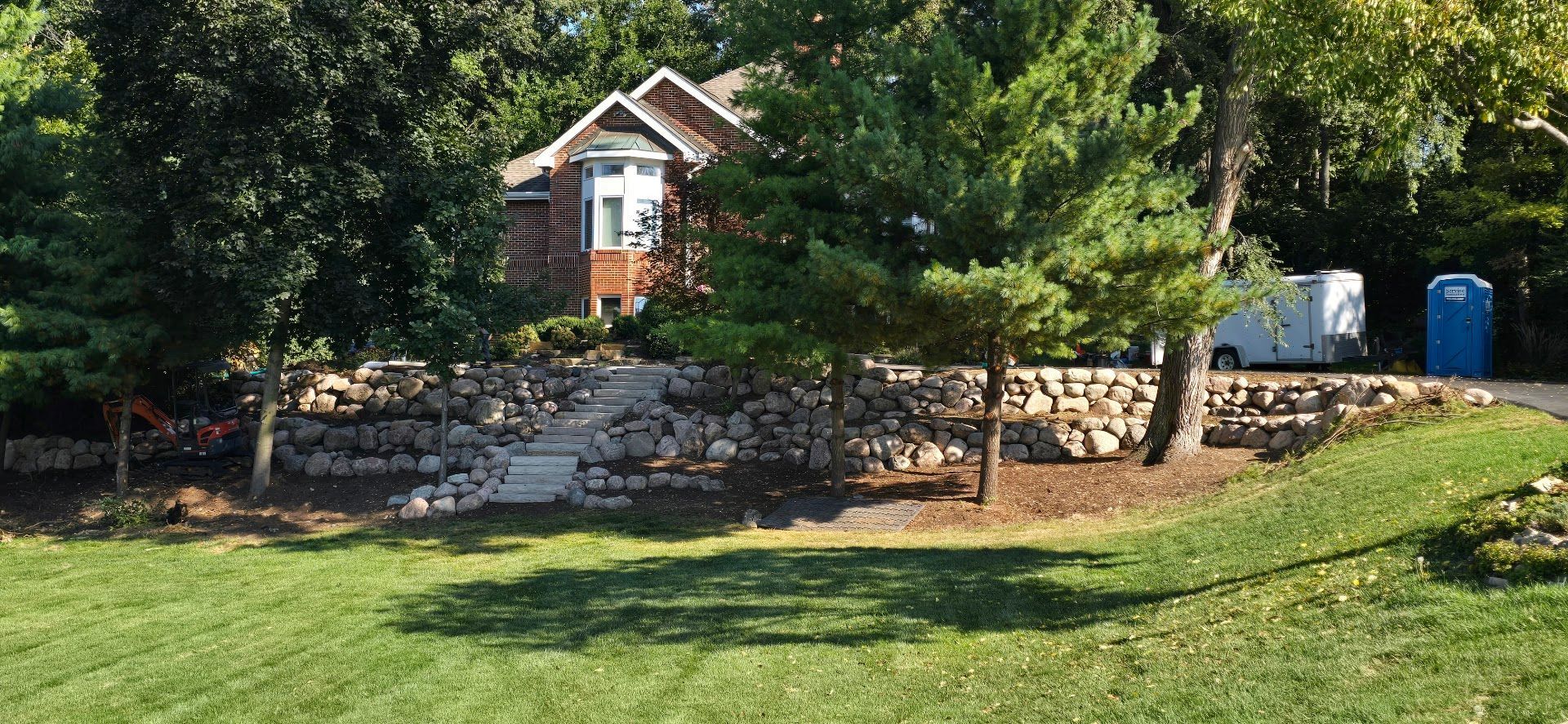 A brick house with a stone retaining wall, stone walkway, trees, and a blue portable toilet on a sunny lawn.