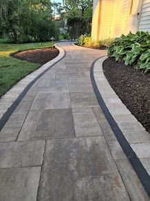 A curved stone paver walkway with a dark border, bordered by mulch and green landscaping next to a building.