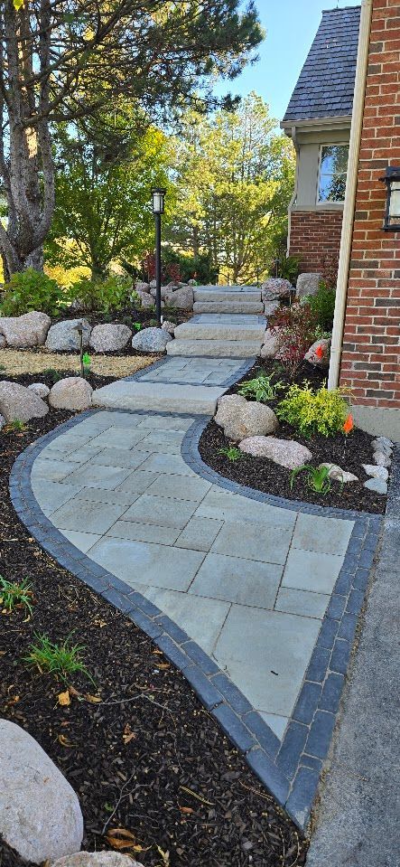 A curving paver walkway with a dark border leads toward stone steps next to a brick house and landscaped garden.