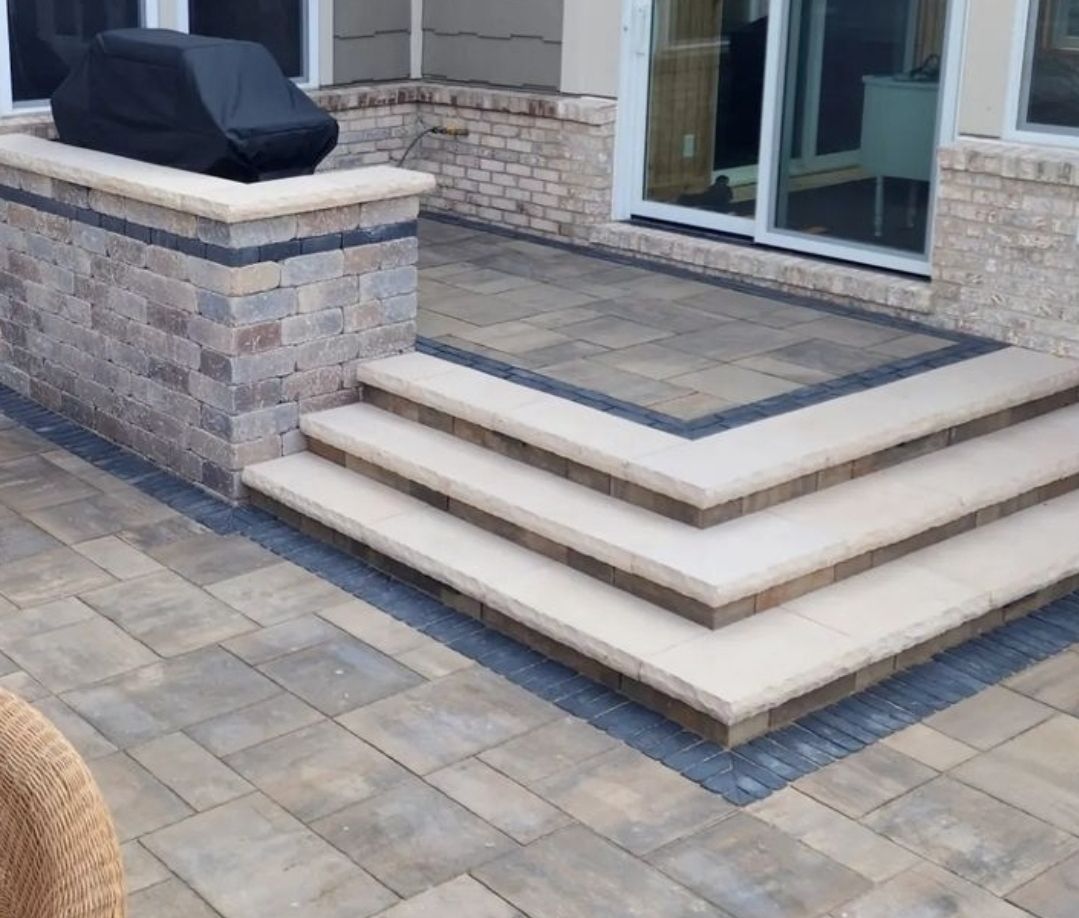 A two-tiered outdoor patio with stone paver flooring, cream-colored steps leading to a sliding door, and a stone grill wall.