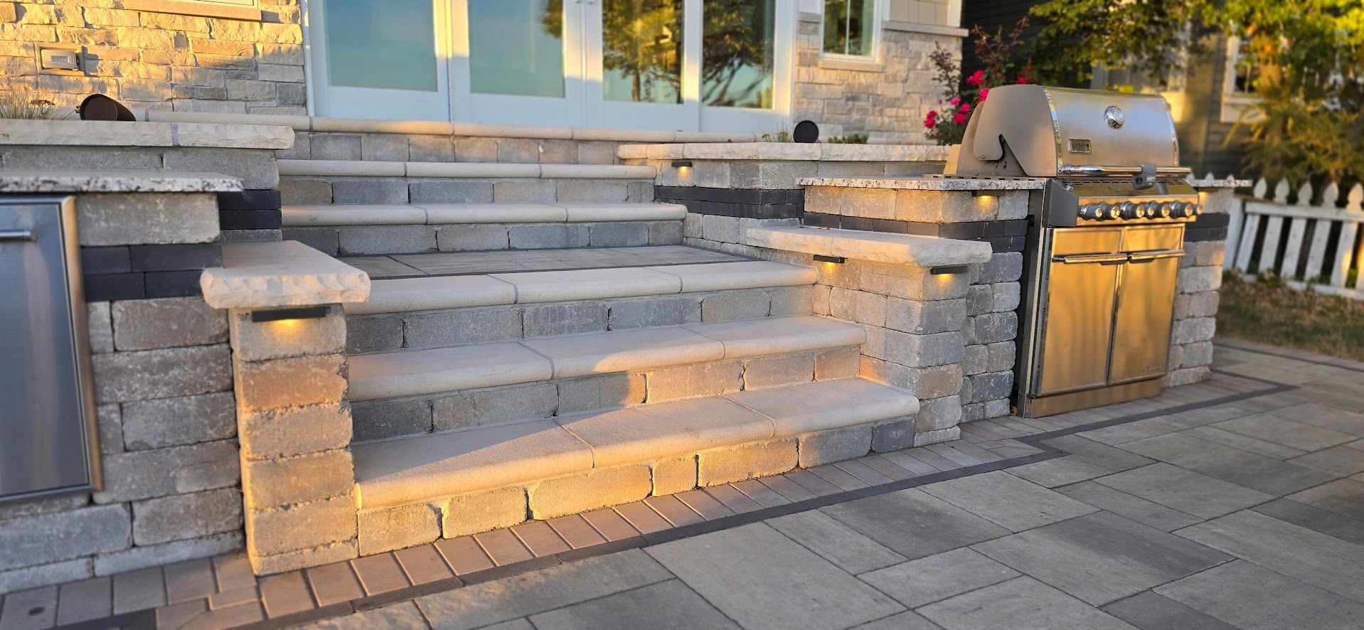 Stone steps leading to a house entrance, flanked by built-in stone pillars, a grill, and a paved outdoor patio area.