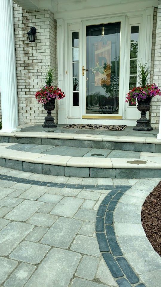 An exterior entrance with a stone porch, steps, and a paved walkway, featuring two potted plants flanking the front door.
