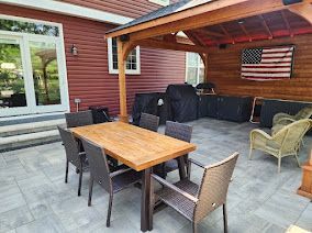 Outdoor patio with a wooden dining set, a covered grill station, and wicker chairs under a wooden pergola at a red house.