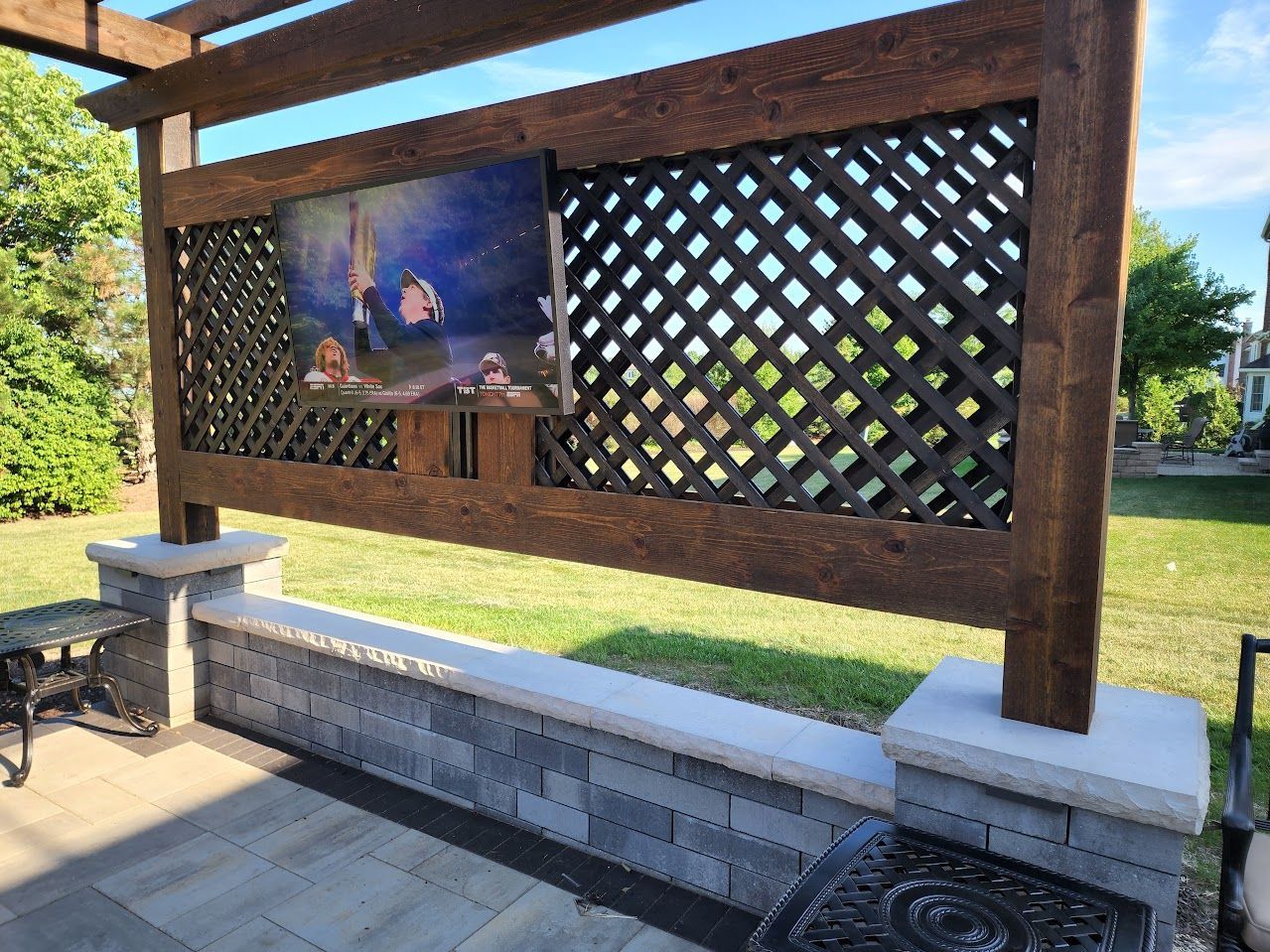 An outdoor television mounted to a wooden lattice wall supported by a gray stone bench in a backyard setting.
