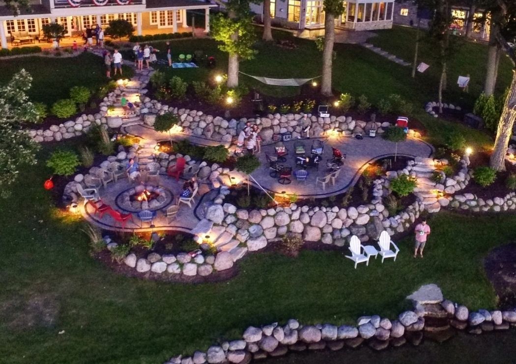 An aerial night view of a landscaped backyard with a stone patio, fire pit area, garden lighting, and a nearby house.