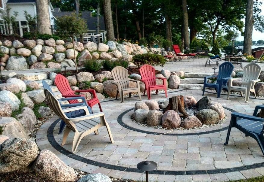 An outdoor stone fire pit patio with several colorful Adirondack chairs, set against a large rock retaining wall.