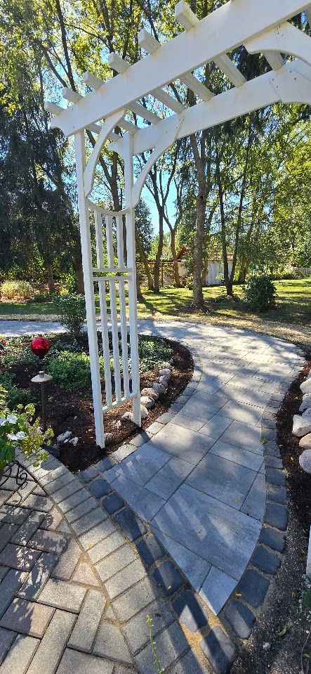 A white wooden garden arbor stands over a stone paver path winding through a landscape with trees and garden beds.