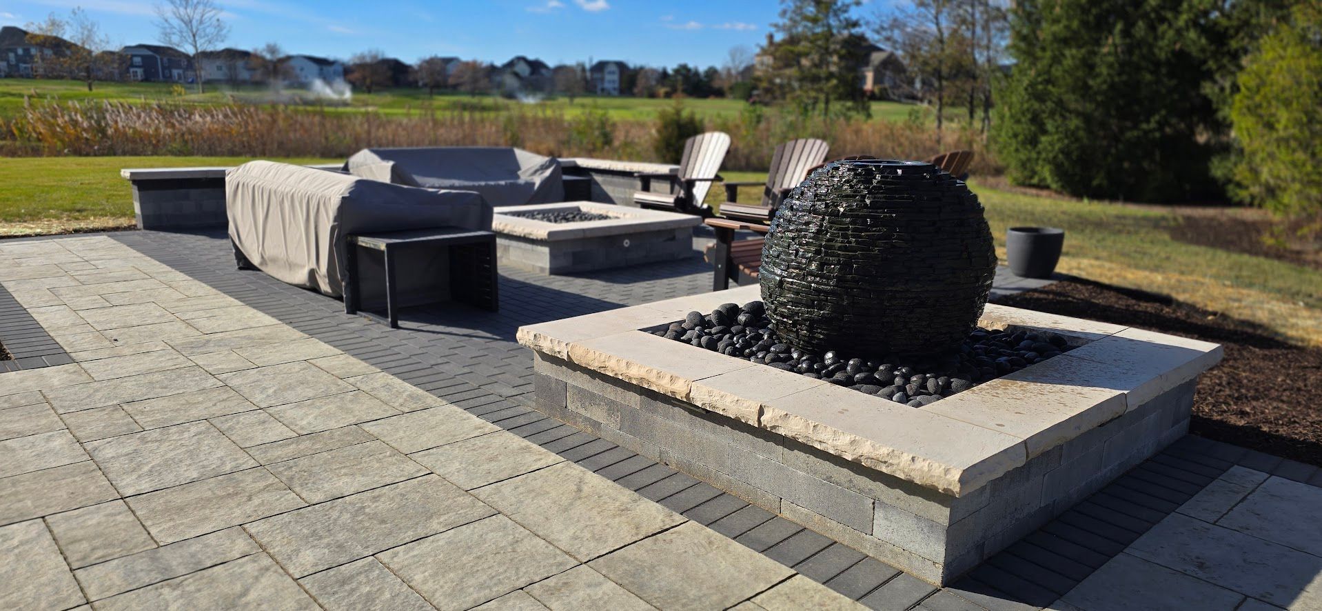 A paved outdoor patio with a large stone water feature, a fire pit, and cushioned seating, overlooking a grassy field.