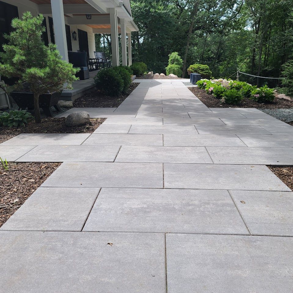 A light gray stone walkway leads to a house porch surrounded by green bushes and trees on a sunny day.
