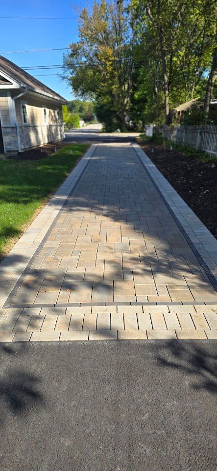 A paved driveway of tan interlocking bricks with grey stone borders, leading past a house and trees to a parking area.