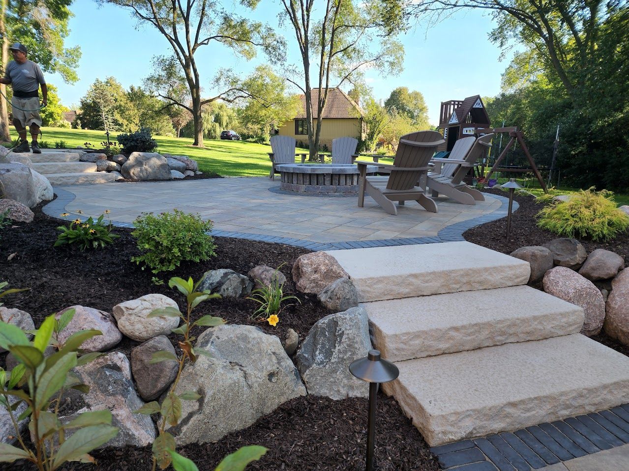 A stone patio with chairs and a fire pit, accessed by three wide steps, surrounded by landscaped boulders and mulch.