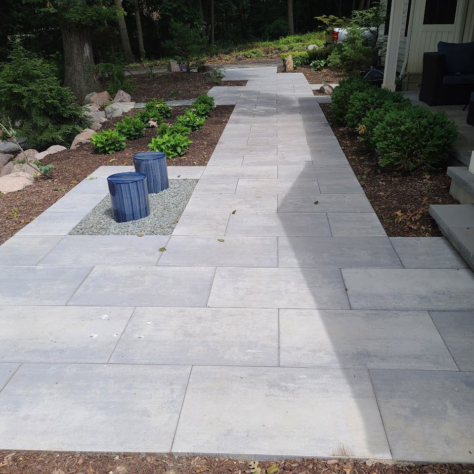 A grey stone patio walkway leading toward a house, flanked by mulch beds, green shrubs, and two decorative blue cylinders.