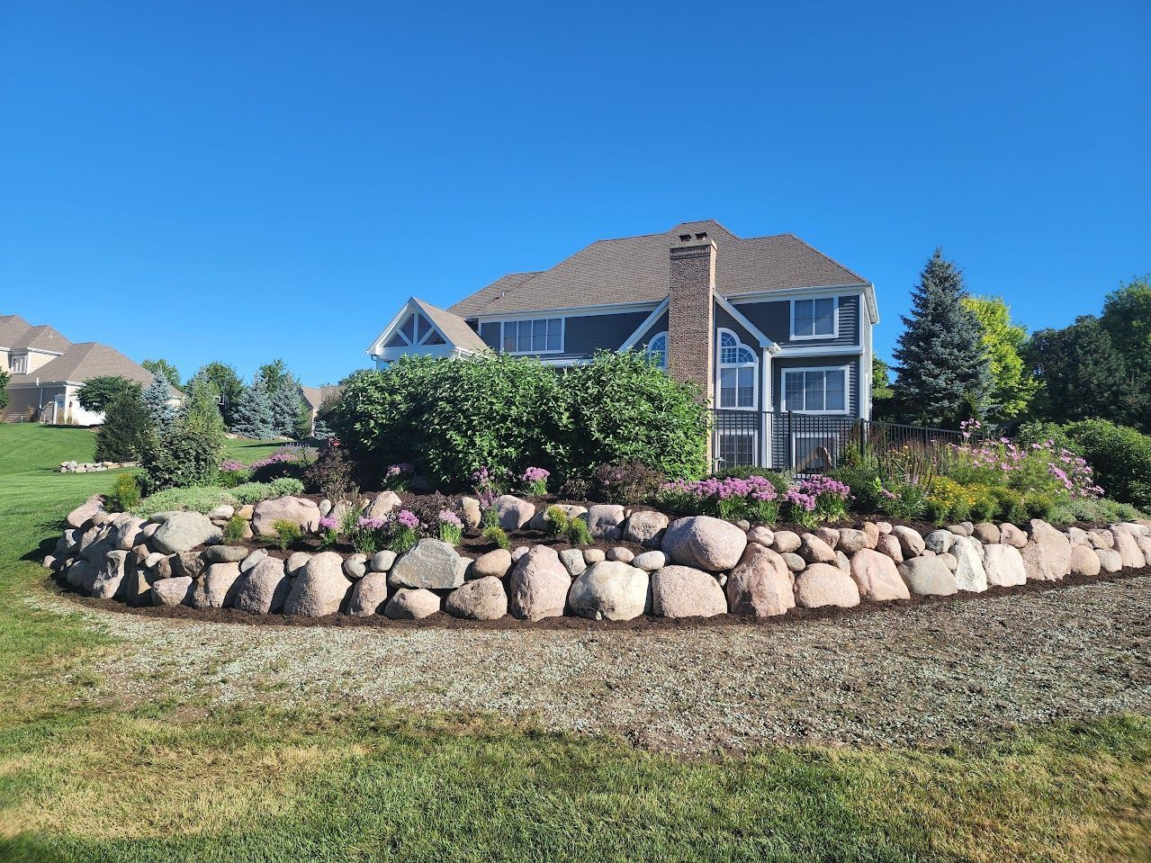 A multi-level backyard landscape featuring a stone retaining wall, lush green shrubs, and flowers in front of a house.