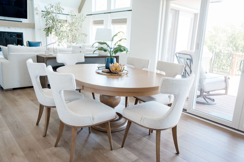 Bright dining room with a round wooden table, white chairs, and sunlight from large windows