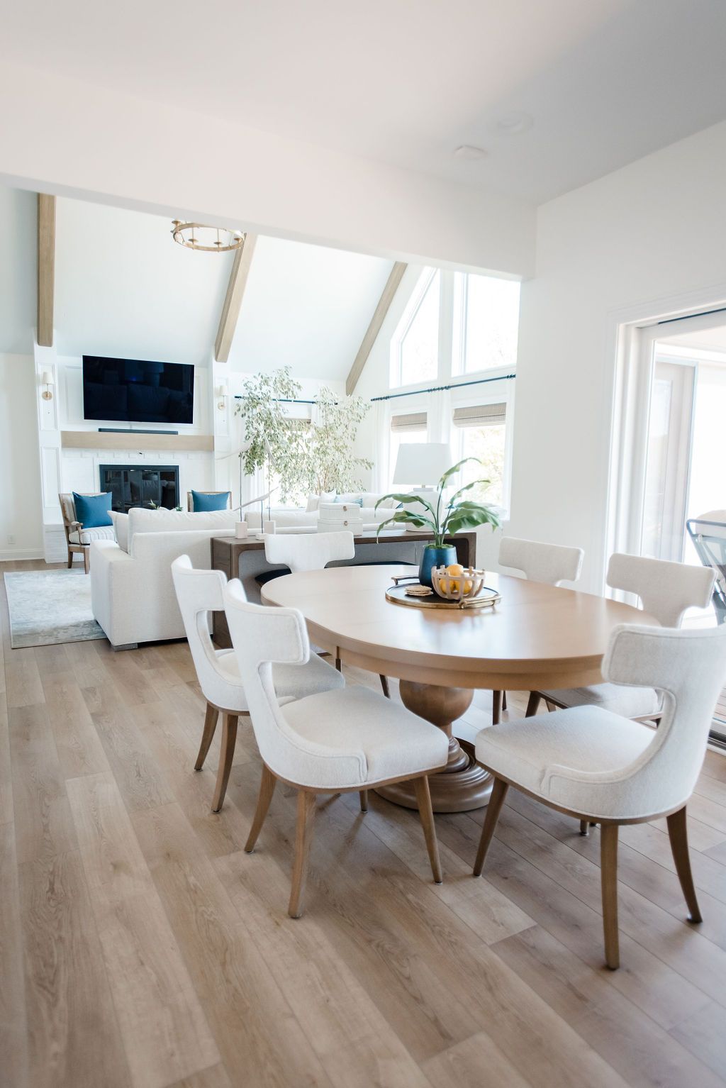 Bright dining room with round wooden table, white chairs, large windows, and fireplace