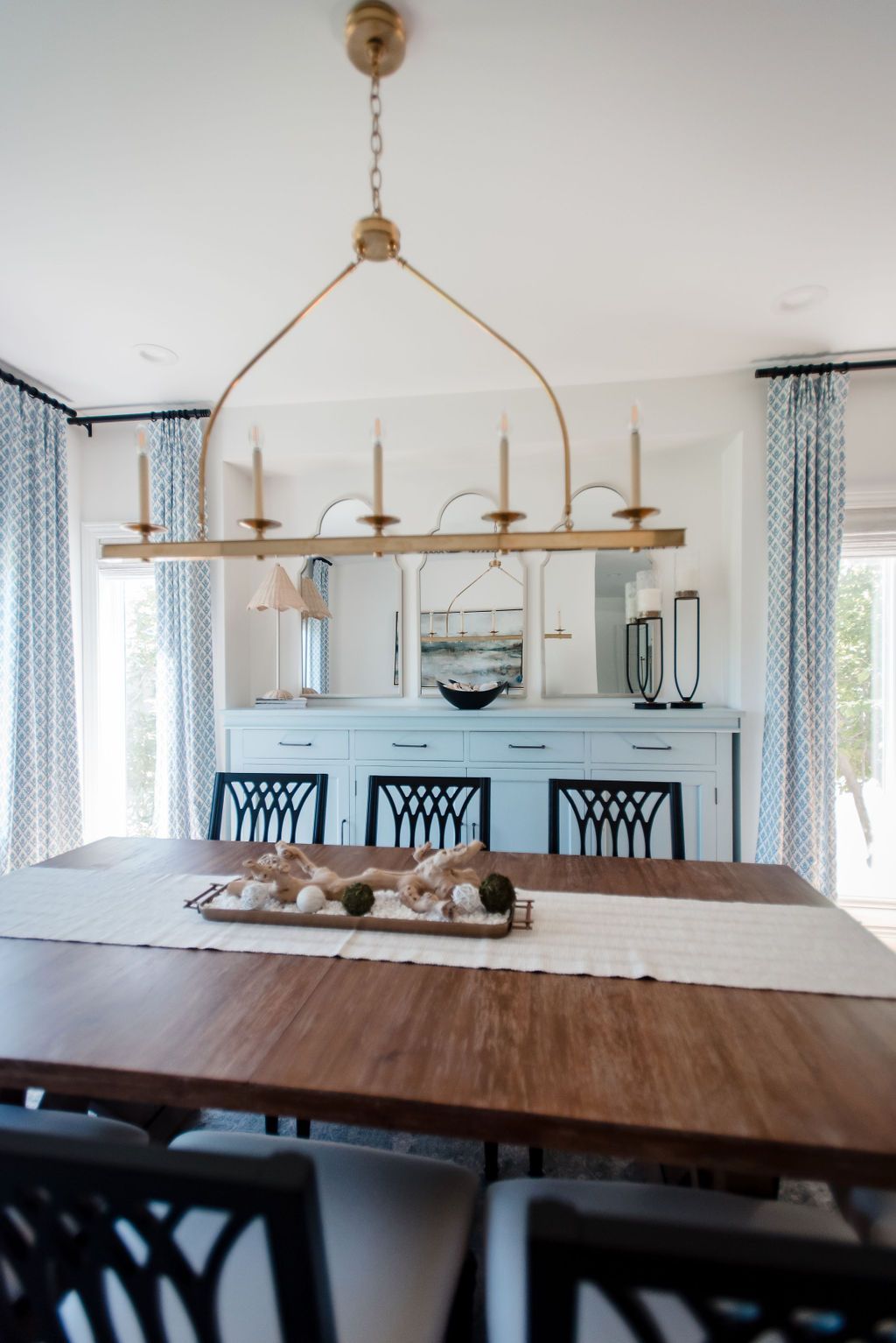 Dining room with a long wooden table, black chairs, and a gold linear chandelier over a pale blue sideboard.