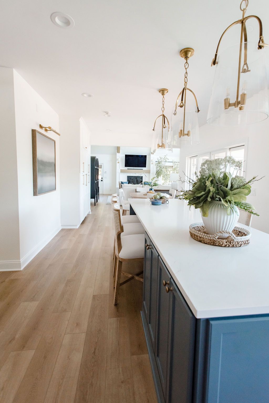 Bright white kitchen with blue island cabinets, wood floors, brass pendant lights, and a potted plant centerpiece.