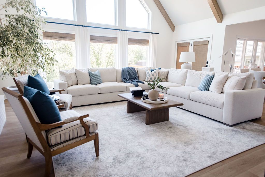 Bright living room with white sofas, blue chairs, and a wooden coffee table under large windows.