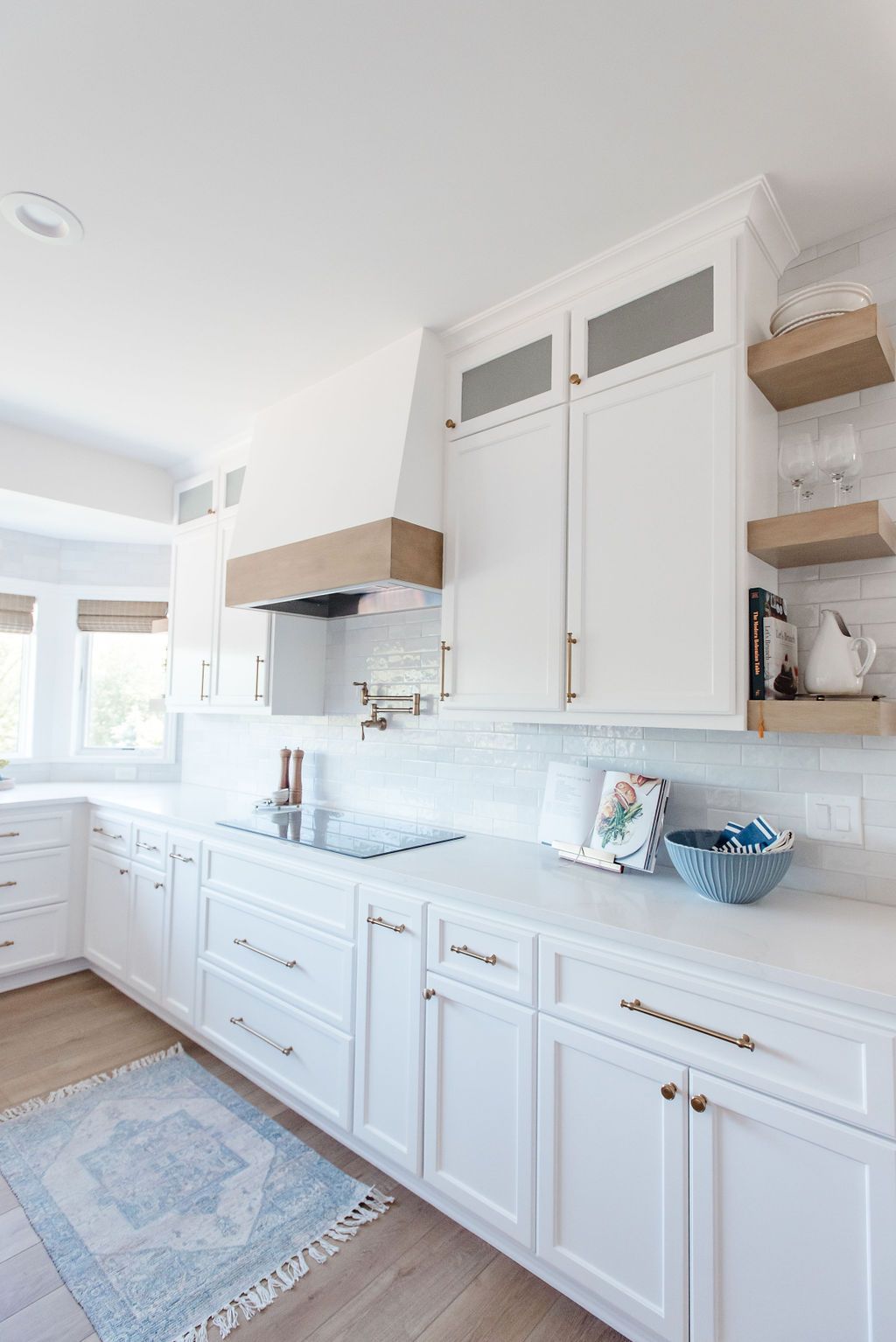 Bright white kitchen with blue rug, marble counters, wood shelves, and tall cabinets.