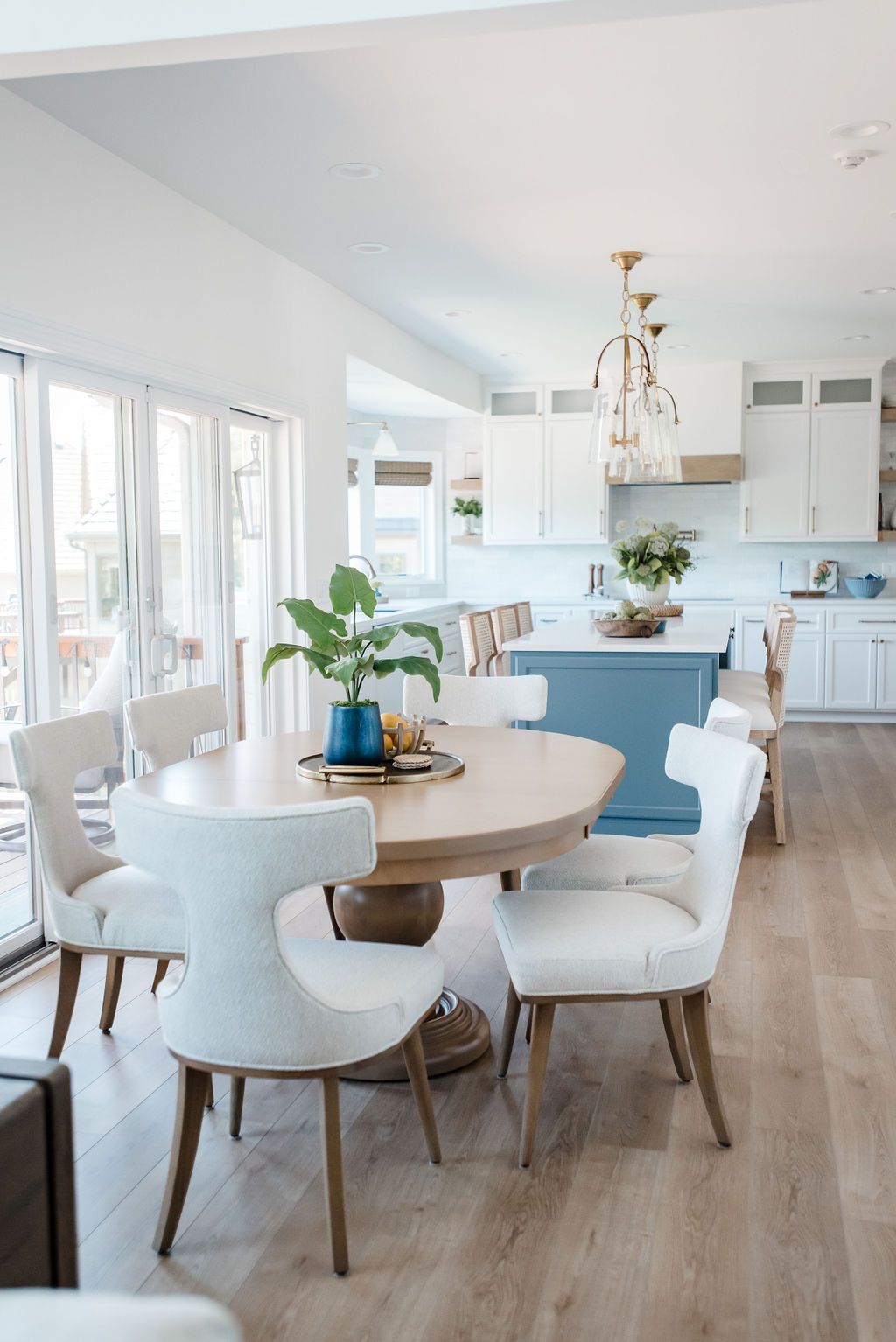 Bright dining room with round wooden table, white chairs, blue bench, and a kitchen in the background