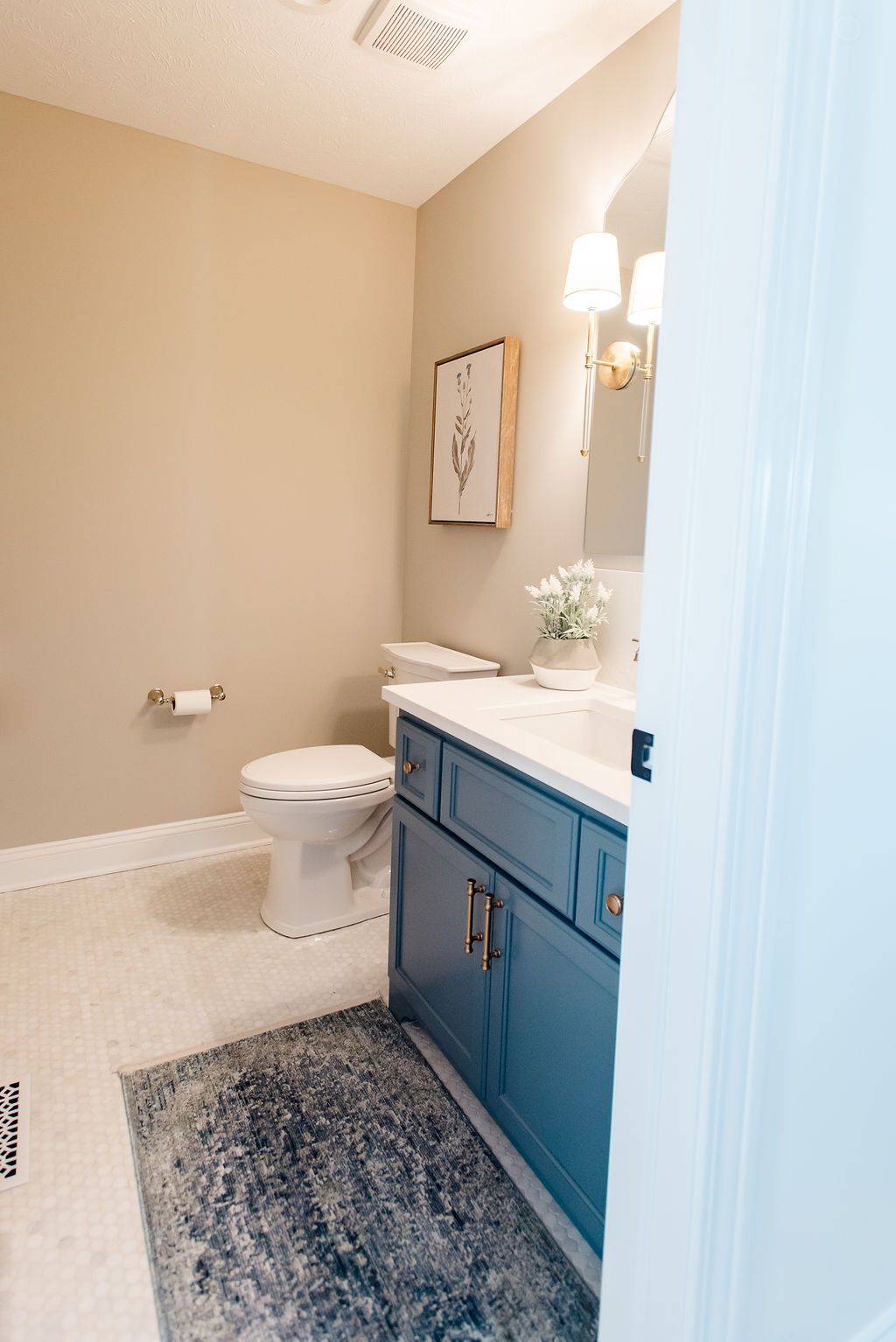 Small beige bathroom with blue vanity, toilet, mirror, wall sconce, and patterned rug