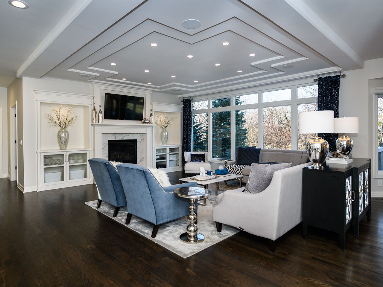 Modern living room with vaulted ceiling, white fireplace, blue chairs, and large windows overlooking trees