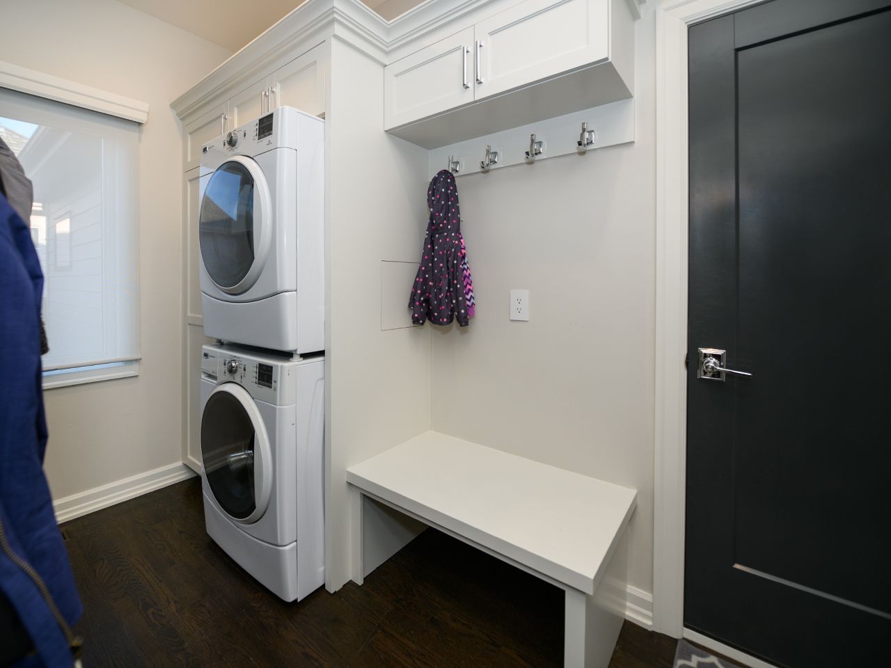 Laundry room with stacked washer and dryer, bench, hooks, and dark door in a white hallway