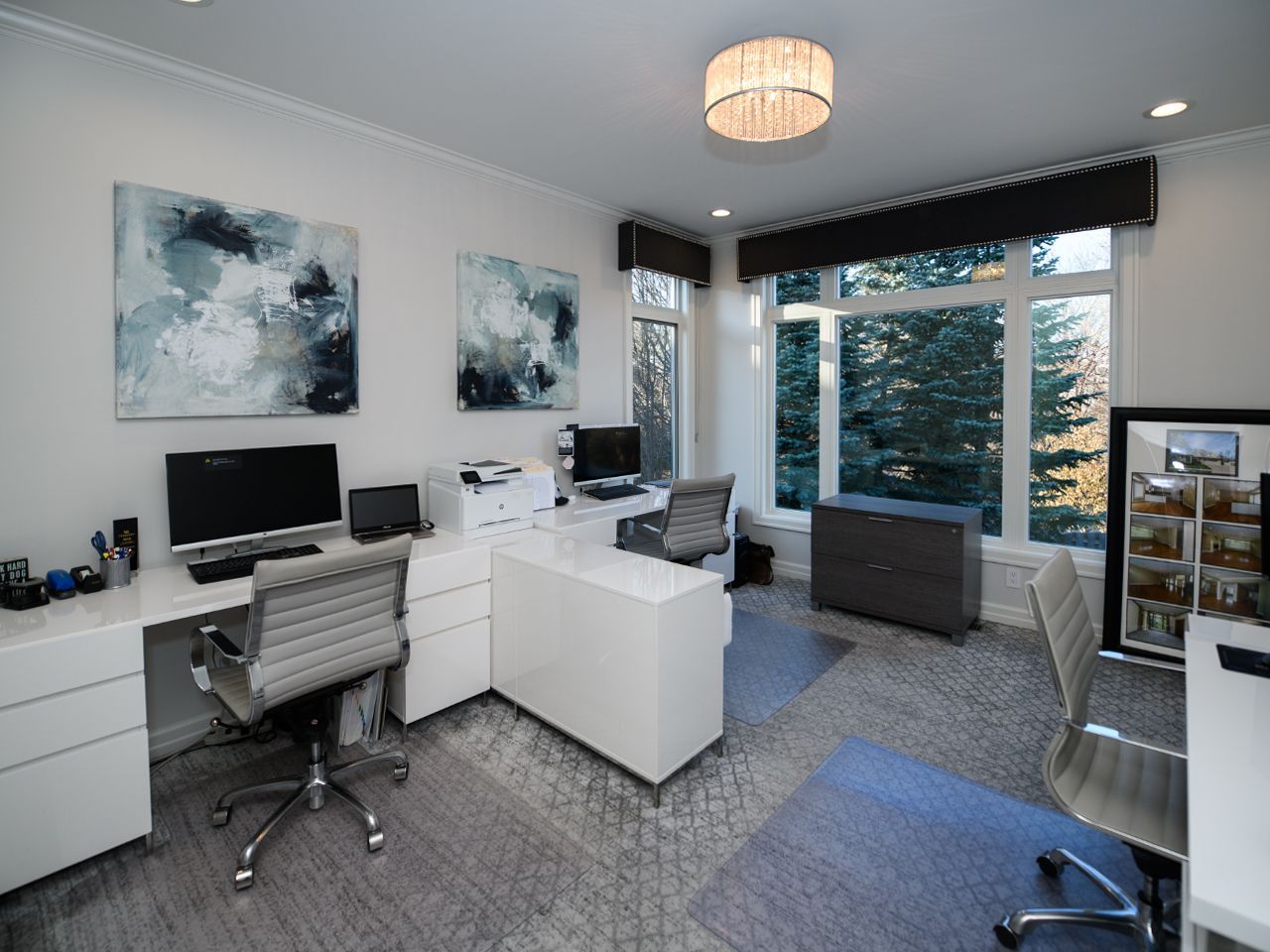 Modern home office with white desk, computer monitors, black chair, and large windows overlooking trees