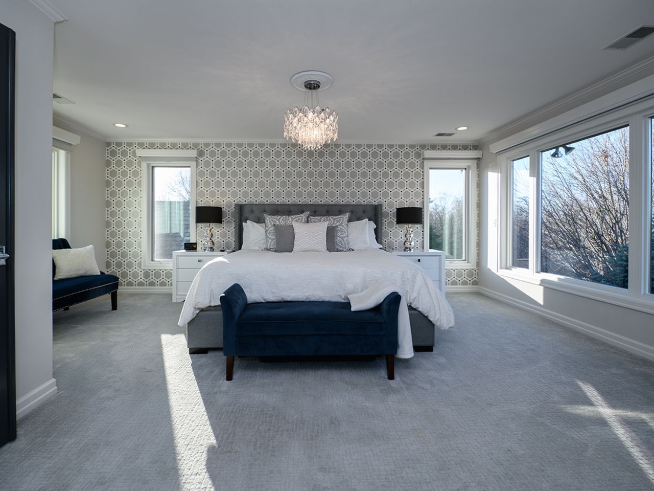 Bright bedroom with a large bed, bench, patterned wallpaper, and floor-to-ceiling windows overlooking trees