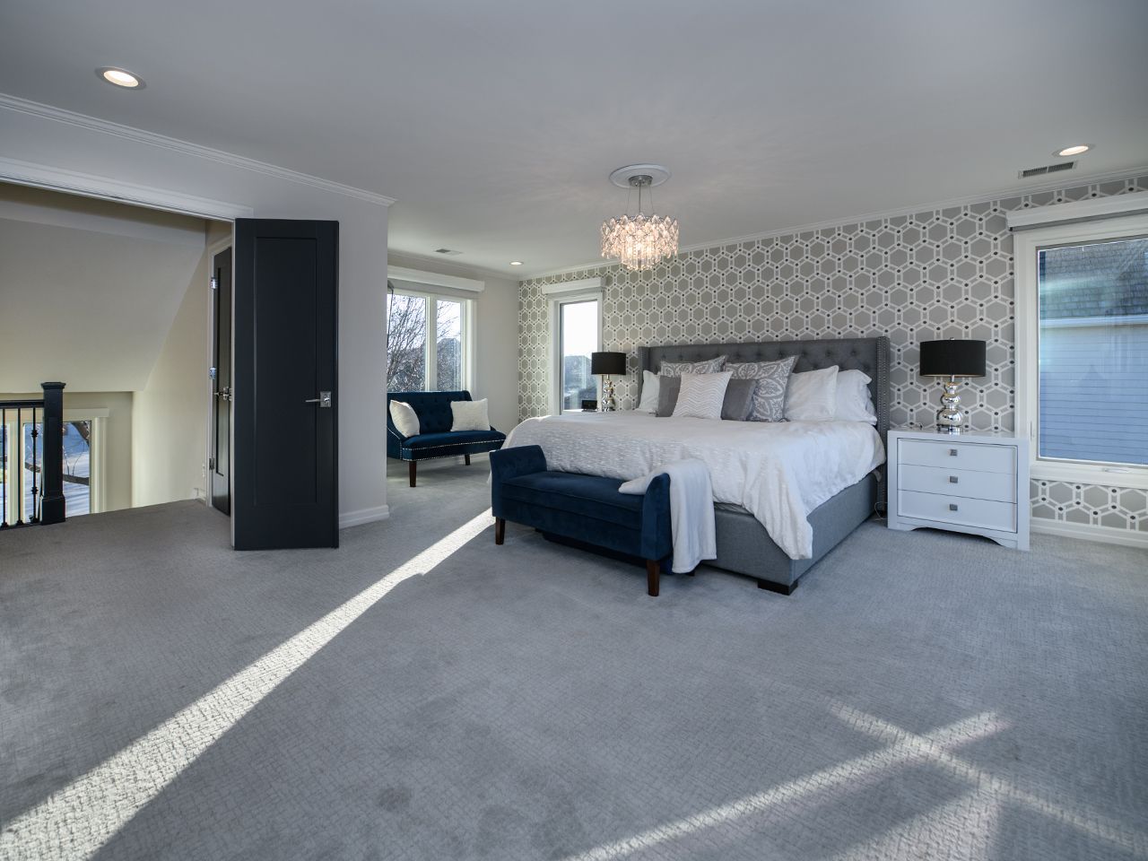 Bright bedroom with a king bed, patterned accent wall, chandelier, and gray carpet in daylight.