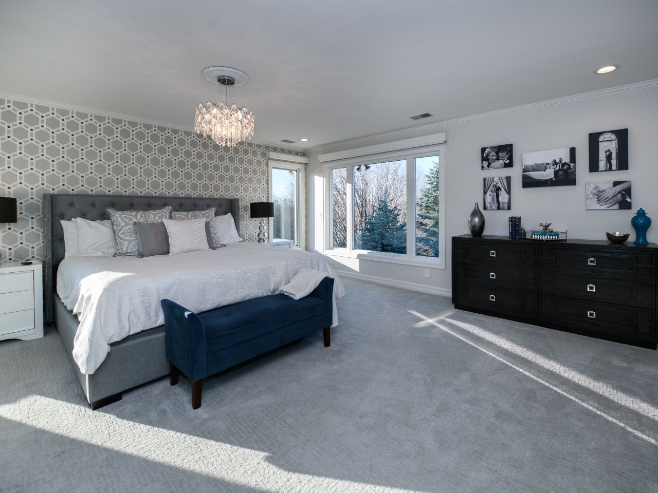 Bright bedroom with gray carpet, white bed, blue bench, patterned wallpaper, and black dresser by a window.