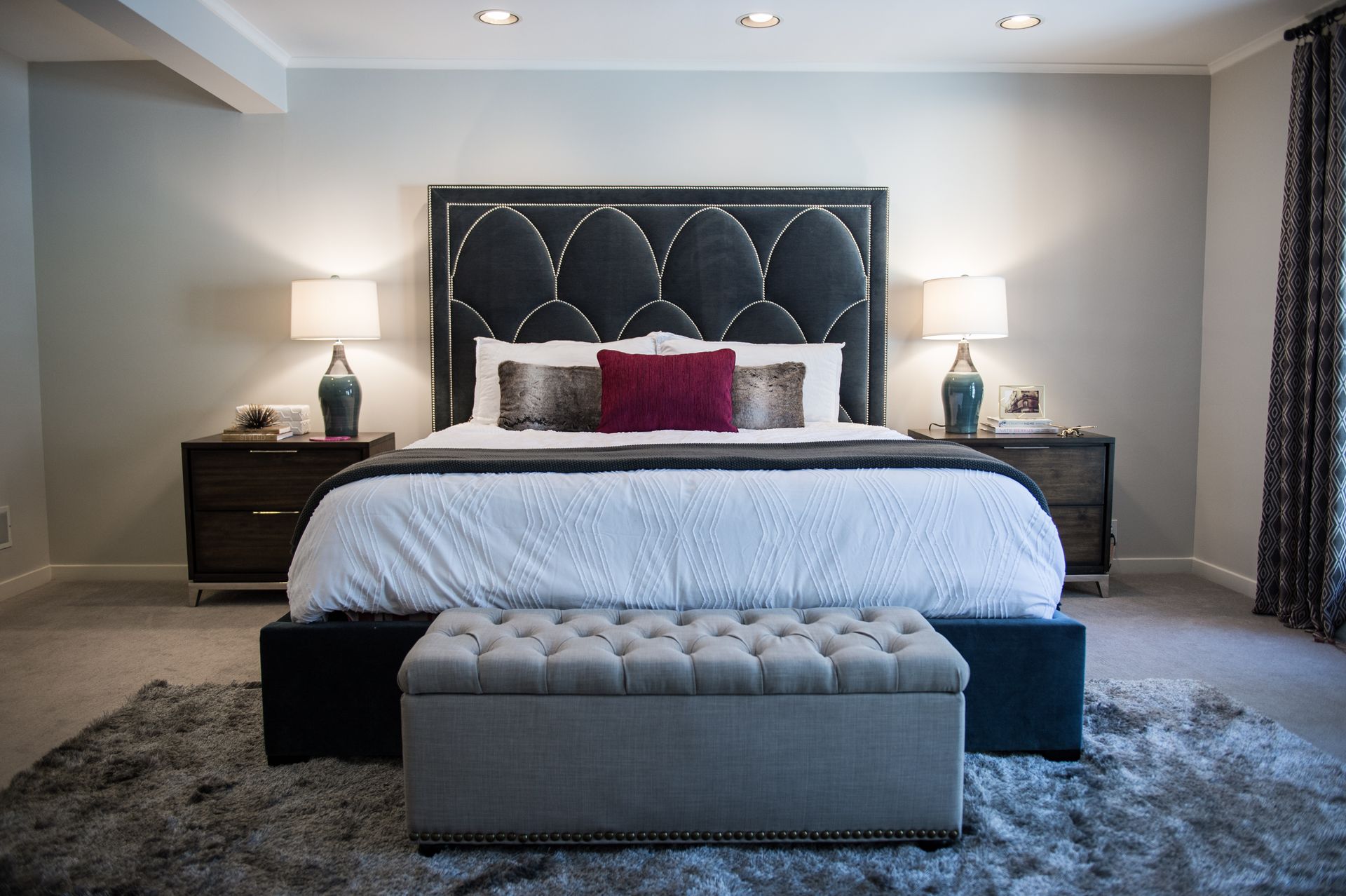 Modern bedroom with a tufted headboard, white bedding, dark nightstands, and a gray bench at the foot of the bed