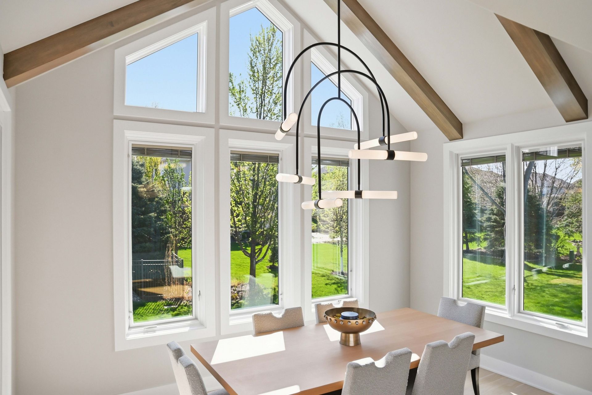 Bright dining nook with large windows, white walls, and a hanging black chandelier overlooking a green garden.