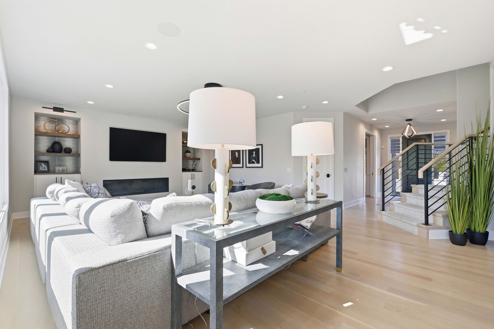 Bright modern living room with gray sectional, wall-mounted TV, glass coffee table, and staircase