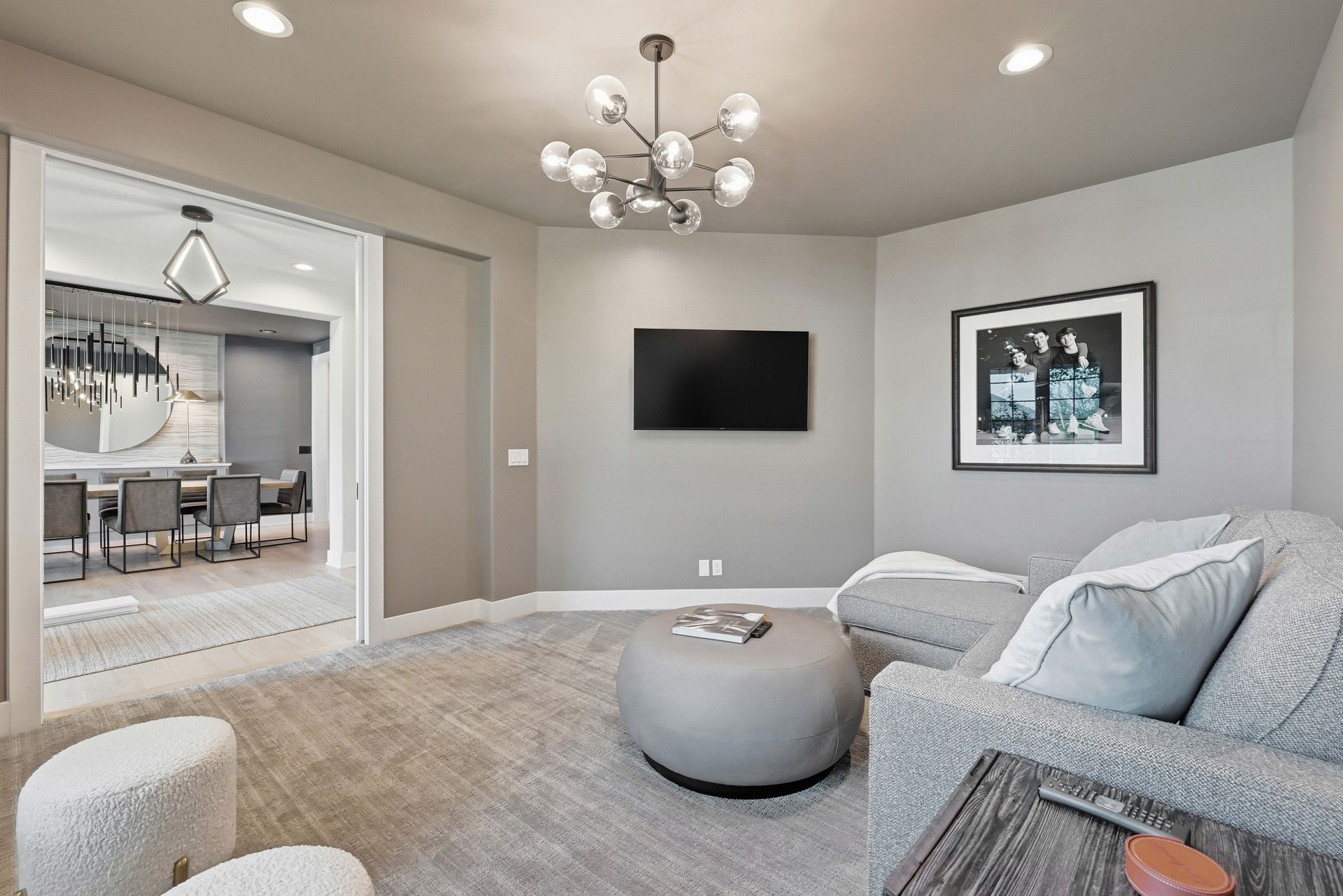 Modern gray living room with wall TV, round ottoman, chandelier, and dining area visible through doorway