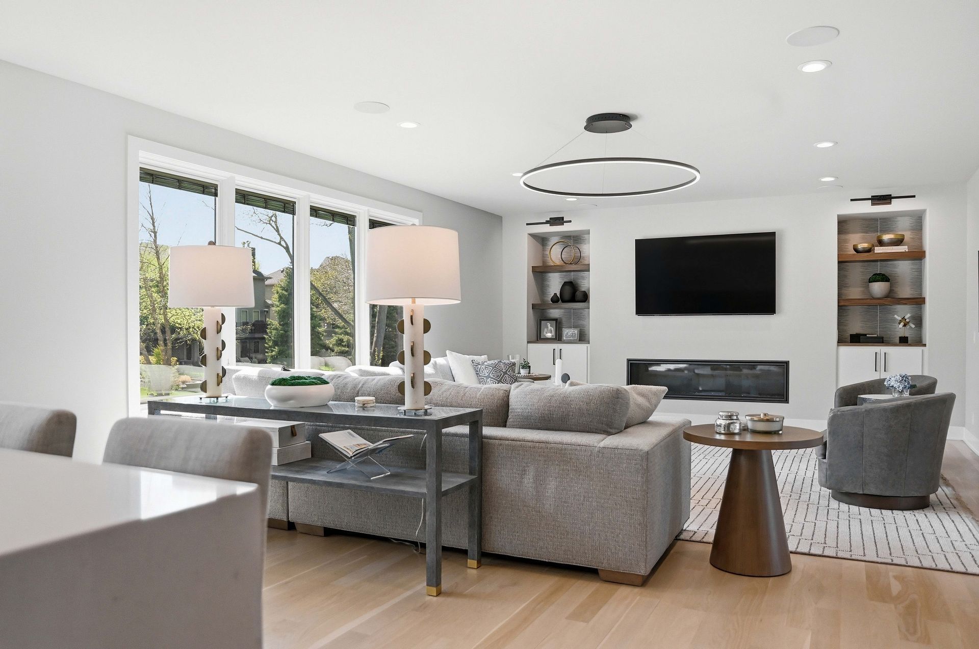 Bright modern living room with gray sofas, wall-mounted TV, round chandelier, and large windows