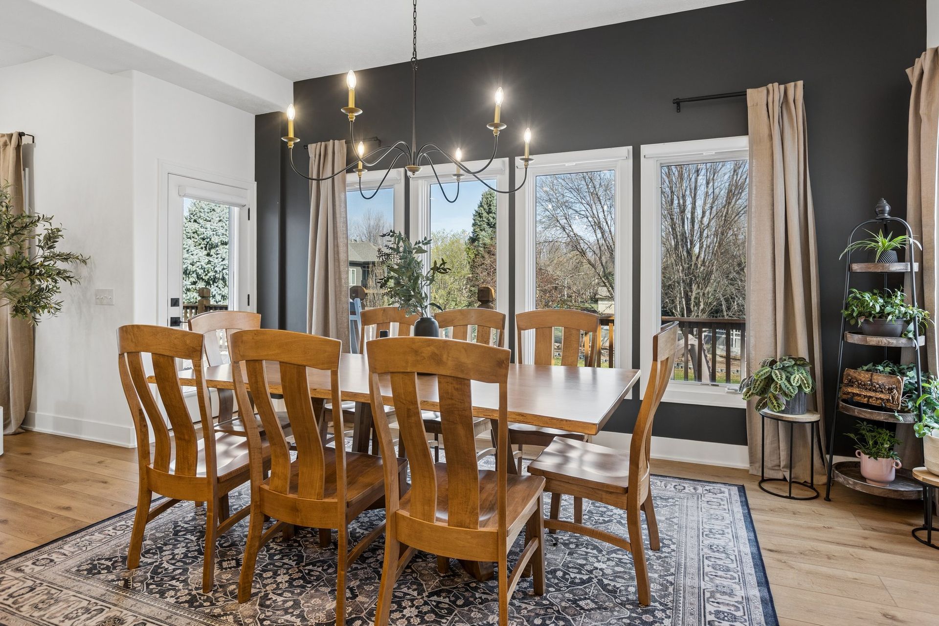 Dining room with wooden table and chairs, dark accent wall, and large windows overlooking trees.