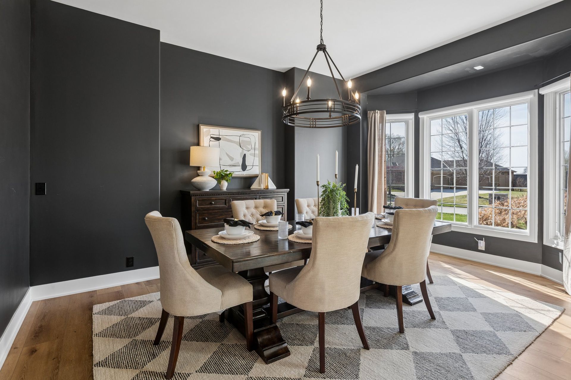 Modern dining room with a round table, beige chairs, dark walls, and large bay windows.